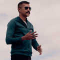 Man wearing a green Athlos Coach Jacket and sunglasses against a cloudy sky