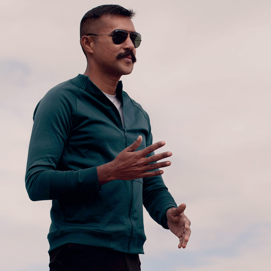 Man wearing a green Athlos Coach Jacket and sunglasses against a cloudy sky